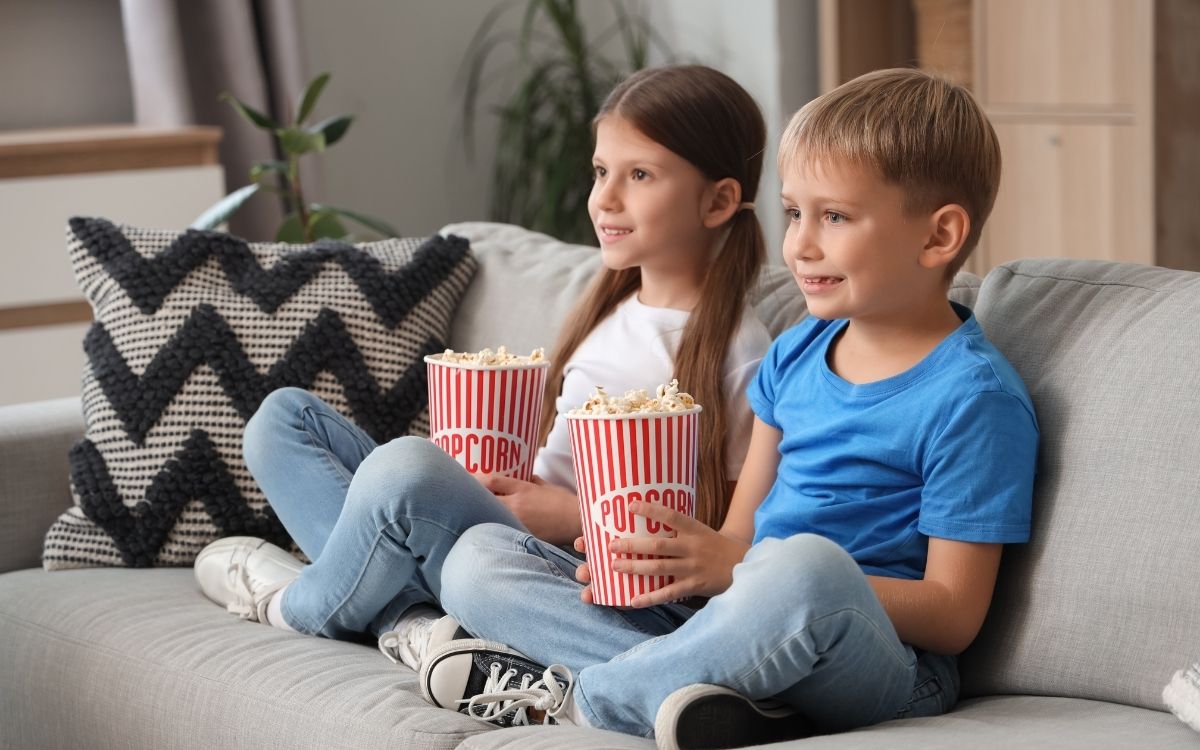 Duas crianças sentadas no sofá assistindo filme e comendo pipoca
